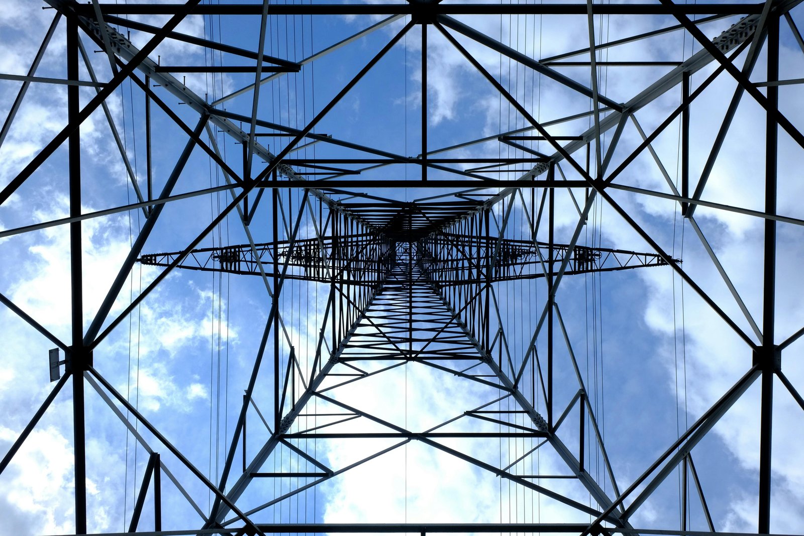 Looking straight up through the interior of a steel lattice electrical transmission tower against a blue sky with white clouds, showing the geometric metal framework and structural engineering of the tower.