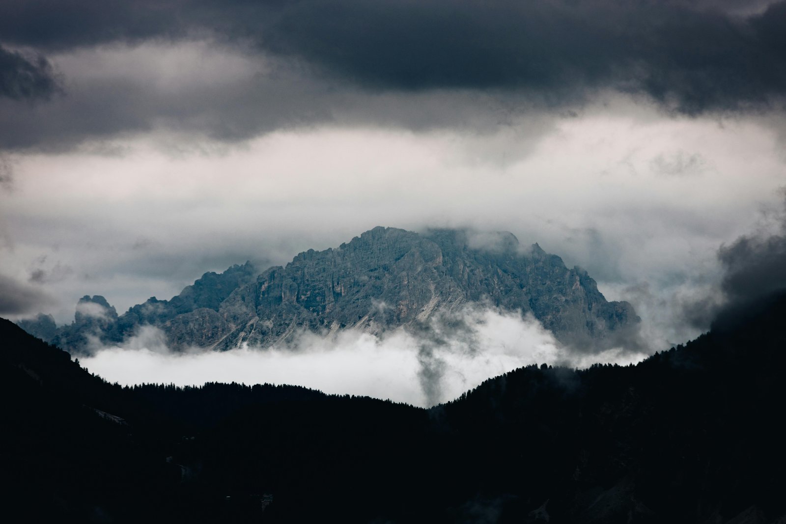 A dramatic mountain valley landscape shrouded in thick storm clouds and mist, with forested foothills in silhouette and a rocky peak partially obscured by swirling clouds under a dark, turbulent sky.