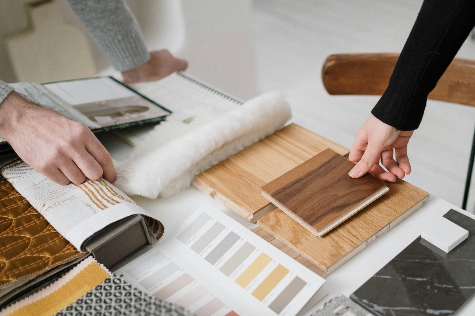 Two people examining and comparing wood flooring samples and material swatches laid out on a white surface, including wood veneer panels, textile samples, and color/finish reference cards.