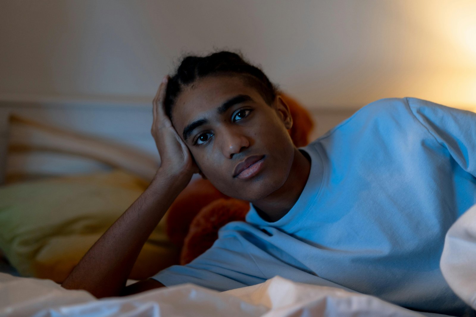 A young person in a light blue shirt lies on a bed in a dimly lit room with warm golden lighting, resting their head on their hand with a contemplative expression.