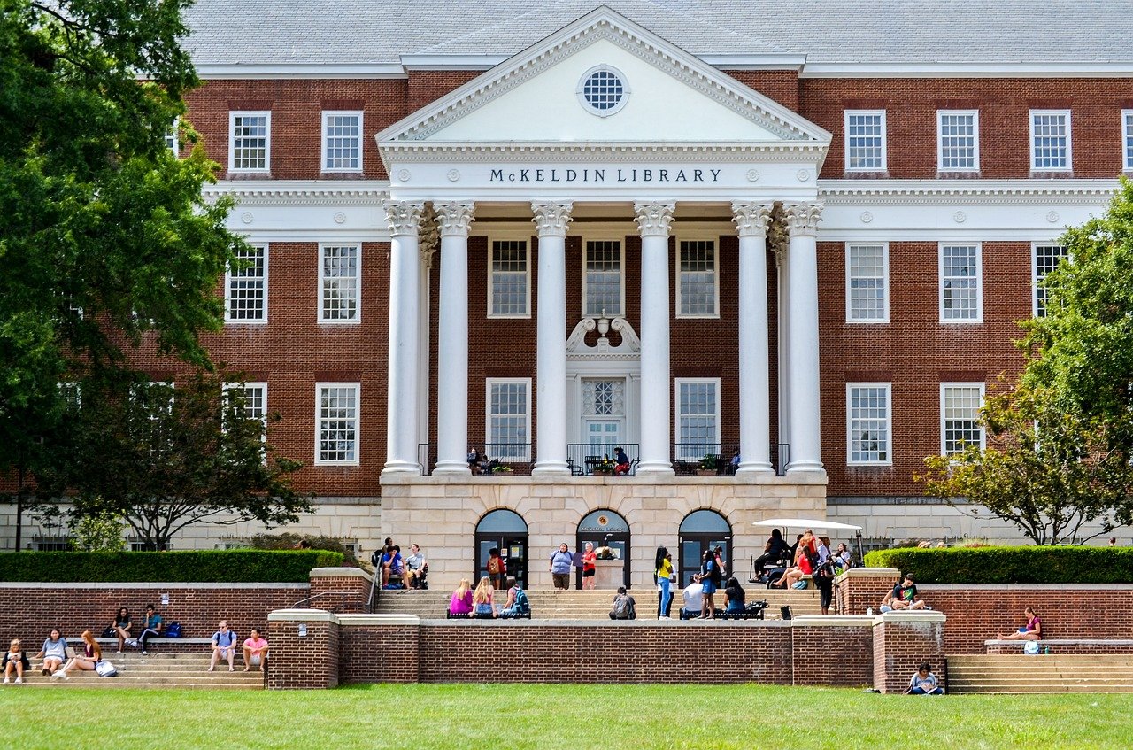 A red brick colonial-style university building with white columns and a peaked roof, surrounded by manicured lawns and mature trees on a clear day.