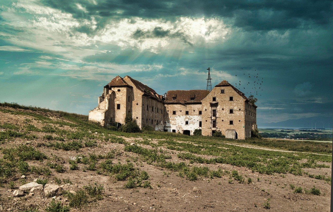 A deteriorated stone building with partial walls and openings stands alone on a barren hilltop under a dramatic cloudy sky, with sparse dry vegetation scattered across the surrounding landscape.