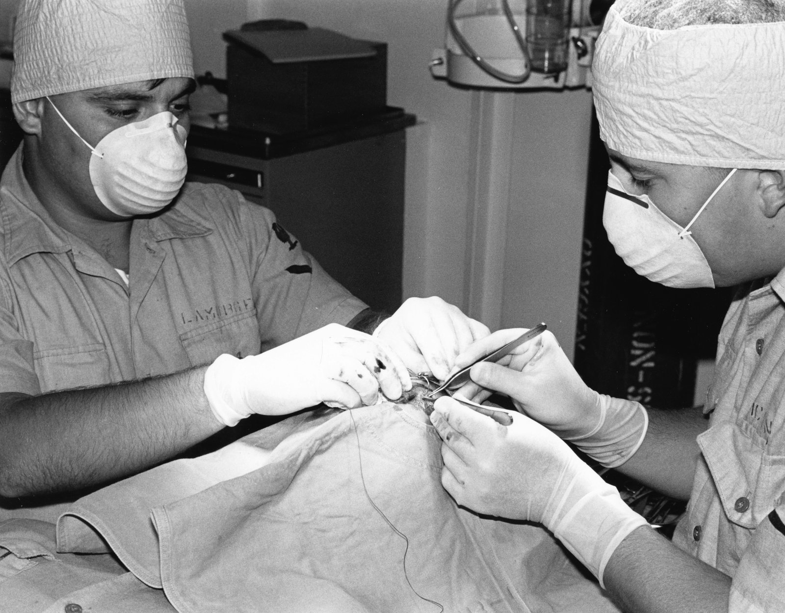 Two surgeons wearing protective masks, head coverings, and gloves perform a surgical procedure on a patient's joint in a sterile operating room environment, with medical equipment visible in the background.