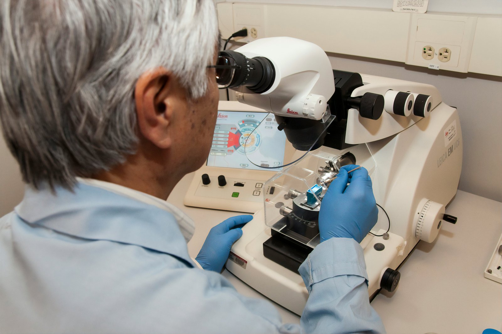 An older scientist wearing a light blue shirt and blue latex gloves examines a specimen under a white binocular microscope in a laboratory setting, with various laboratory equipment and instruments visible on the workbench.