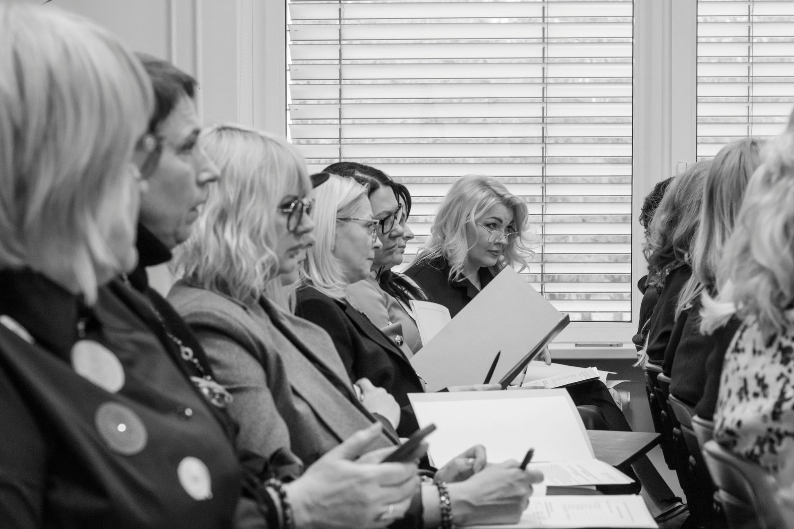 A black and white photograph of a group of women seated around a table in a meeting room with louvered windows in the background, holding papers and documents while engaged in discussion.