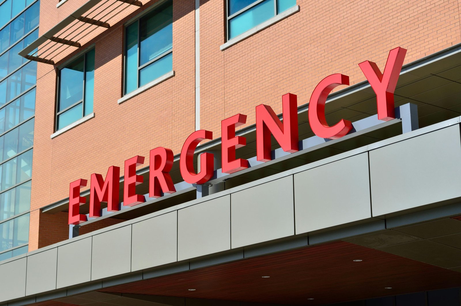 A large red "EMERGENCY" sign mounted on the exterior facade of a modern brick hospital building with glass windows and an overhang structure.