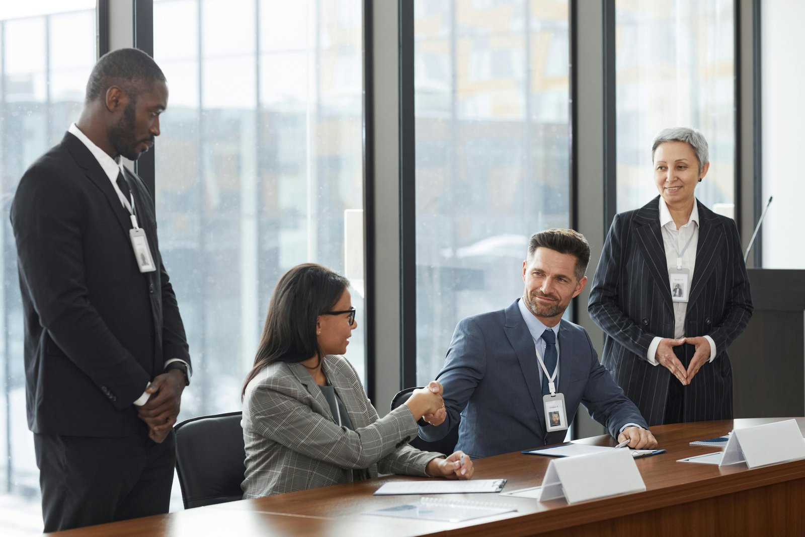Four business professionals in formal attire gathered around a wooden conference table in a high-rise office with floor-to-ceiling windows overlooking a cityscape; two seated and two standing, appearing to discuss documents or agreements in a corporate meeting.