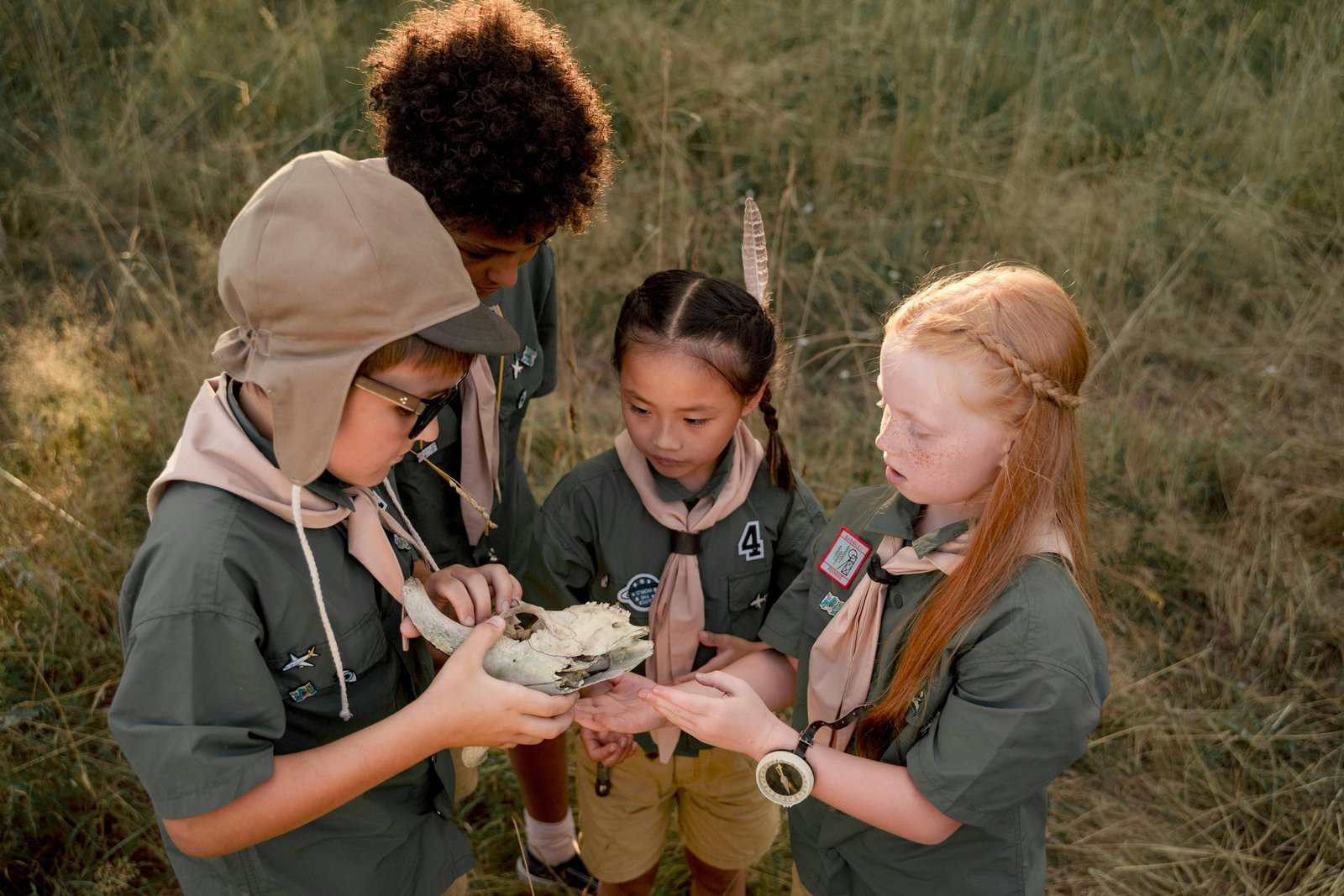 Four young students in scout or outdoor education uniforms examine a small object or specimen together while standing in a woodland area with dry grass and trees in the background.