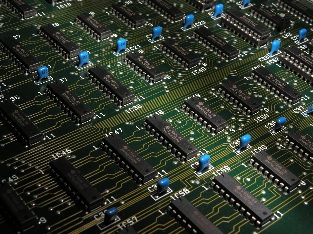 A close-up photograph of an illuminated green circuit board with blue and cyan glowing electronic components and microchip pathways visible against a dark background.