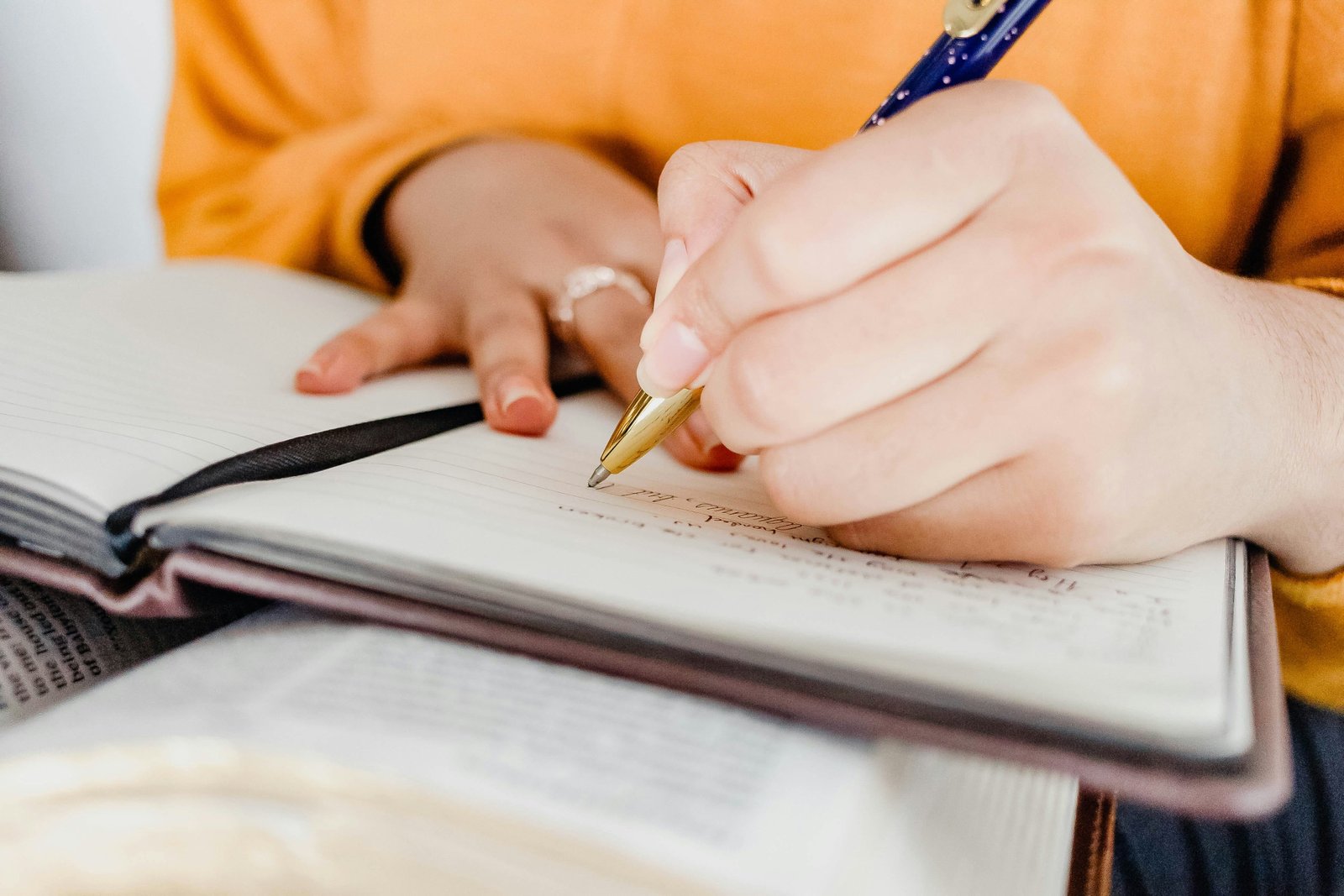 A person wearing a yellow/orange shirt writes in an open notebook on a desk with a pen, with documents and papers visible nearby.